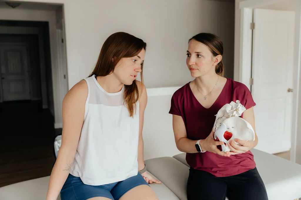 Dr. Danaya Kauwe sitting with a patient in her home, explaining pelvic anatomy using an anatomical model during a postpartum consultation